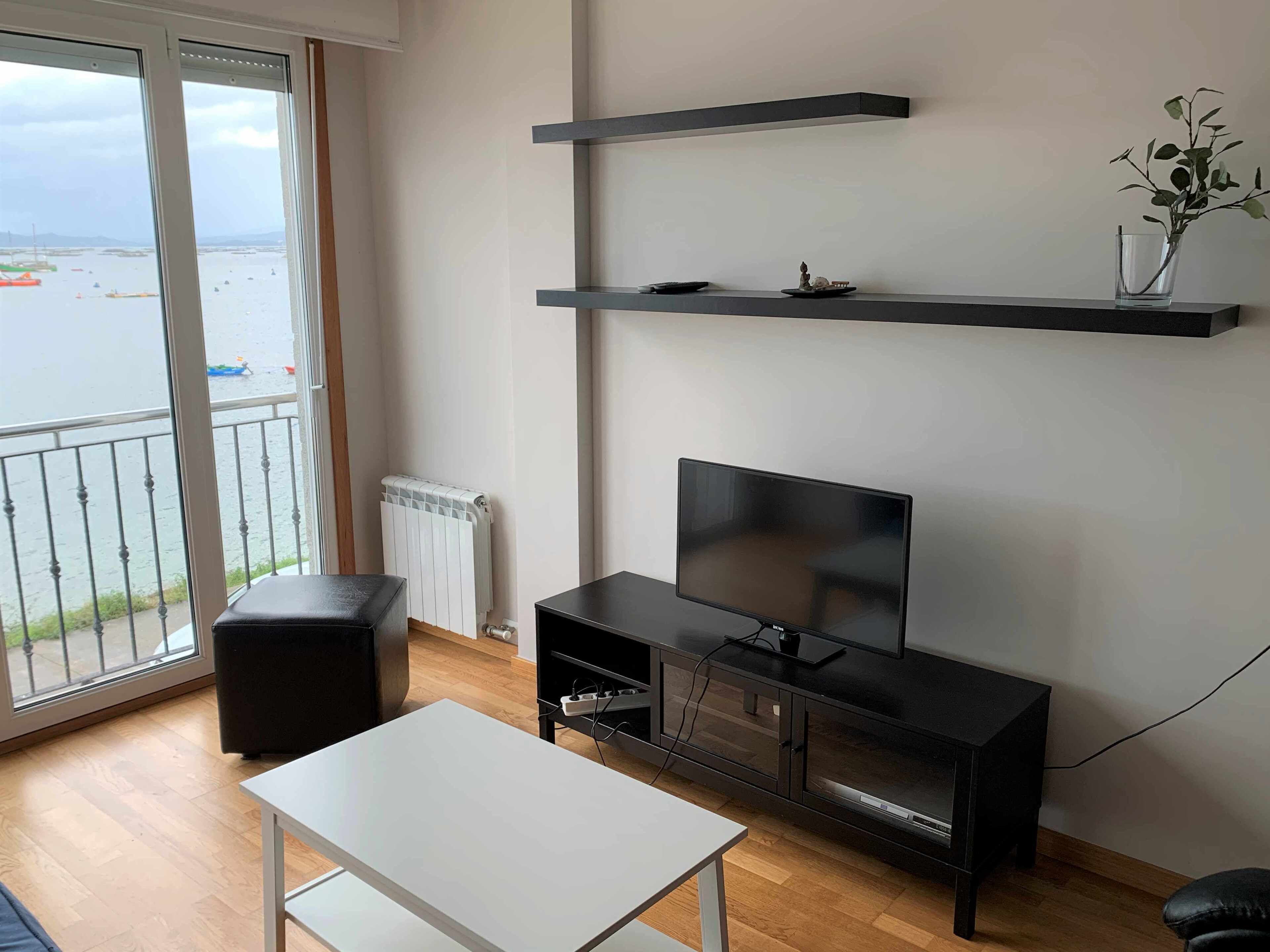 A modern living room with a TV on a black stand, a white table, and a balcony with a view of boats.