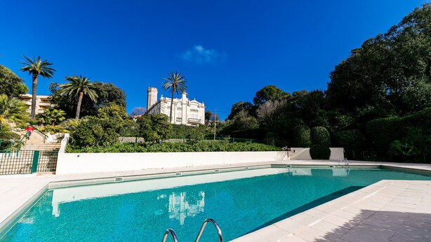 A swimming pool with clear blue water, surrounded by a white deck and lush greenery. In the background, there is a large building with a tower. - LE CHATEAU DE LA PINEDE AP4198 (Juan-les-Pins)