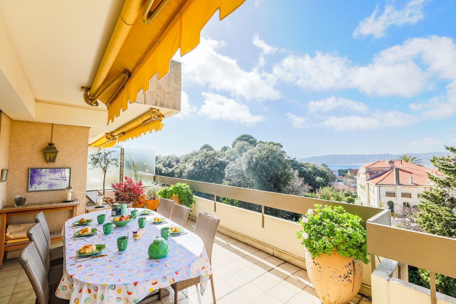 A balcony with a table set for dining, surrounded by wicker chairs, and overlooking a scenic view of trees and buildings.