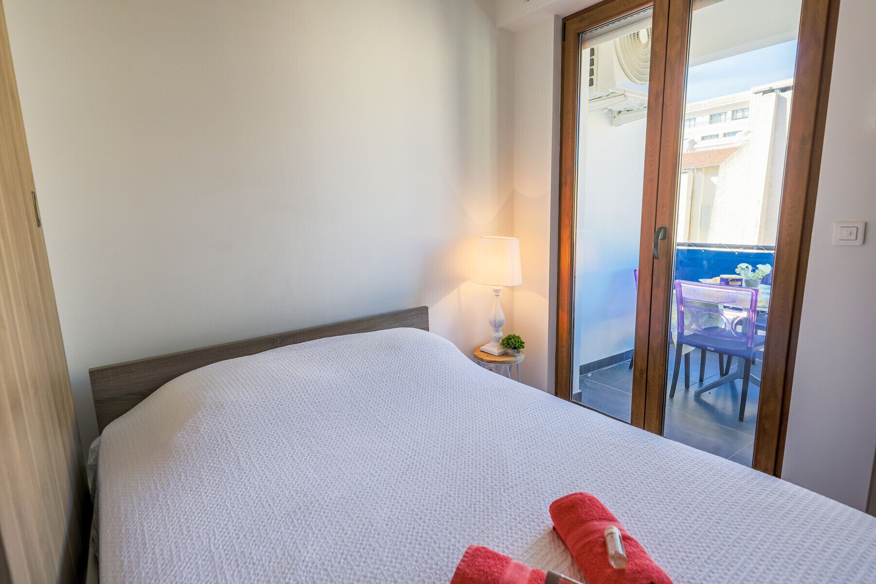 A hotel room with a bed, a nightstand with a lamp, and a view of the outside balcony.