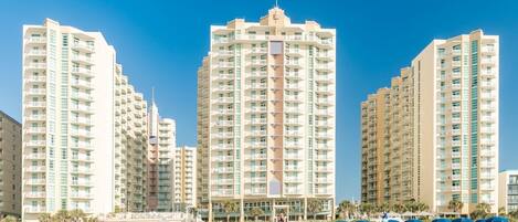 Un hotel frente a la playa con varios edificios altos, sombrillas y un cielo azul despejado.