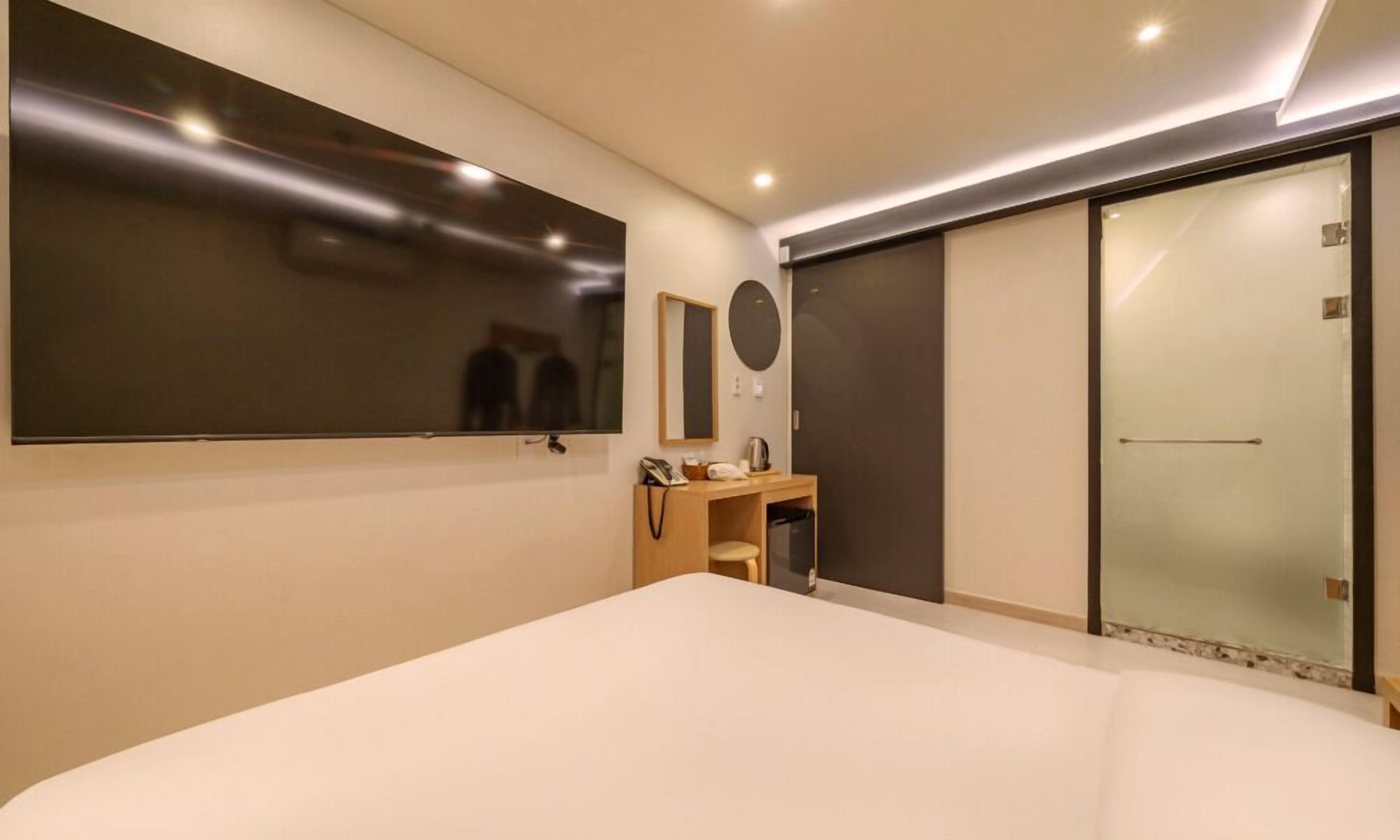 A modern hotel room with a large bed, a vanity with a mirror, a small desk, and a sliding door closet.