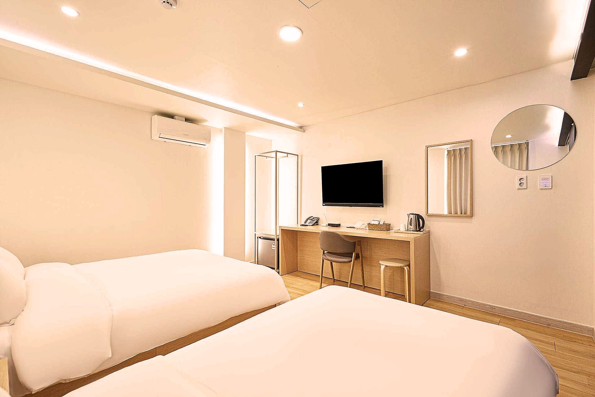 A hotel room with two beds, a desk with a chair, a television, a mirror, and an air conditioning unit.