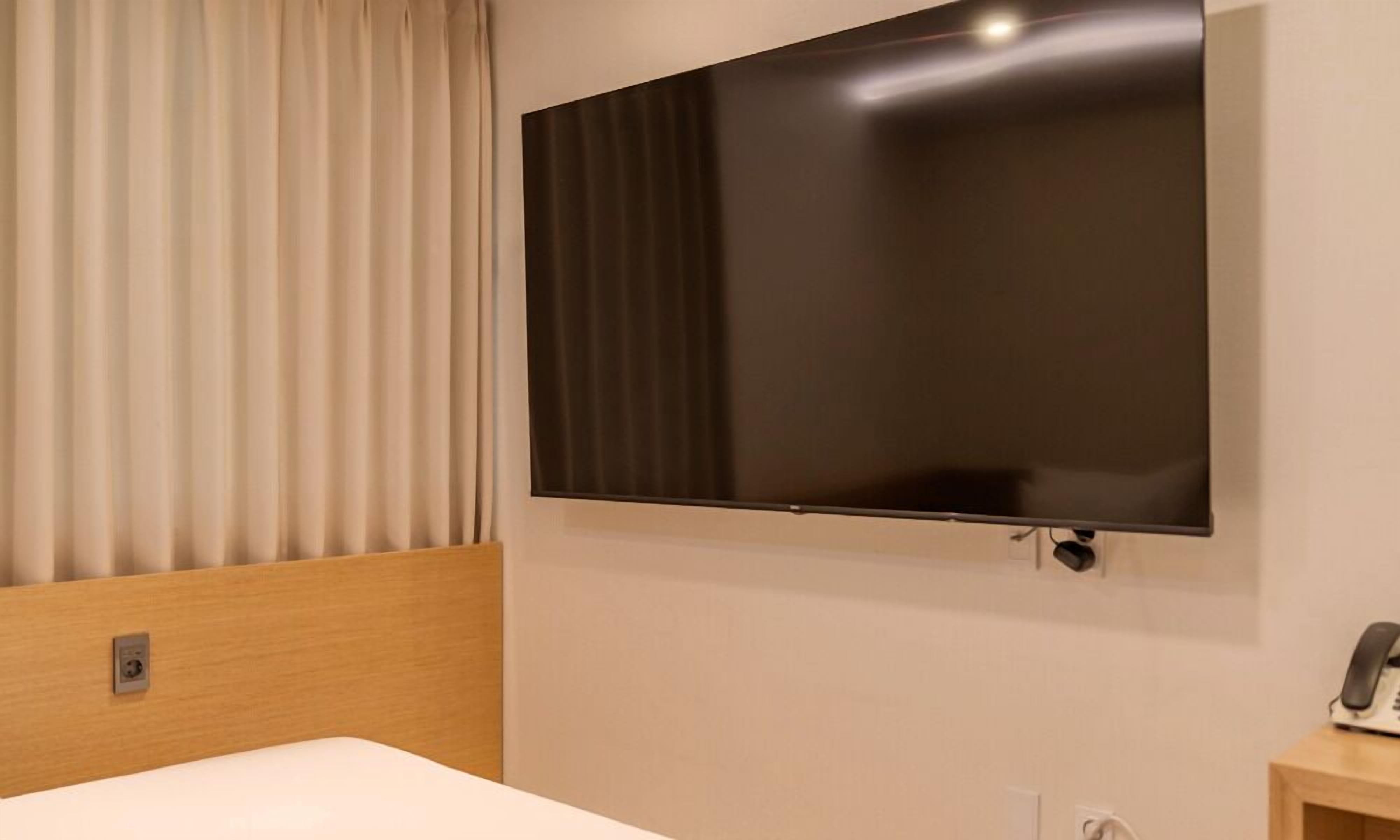 A hotel room with a large flat-screen TV mounted on the wall, a wooden headboard, a white bed, and a bedside table with a telephone.