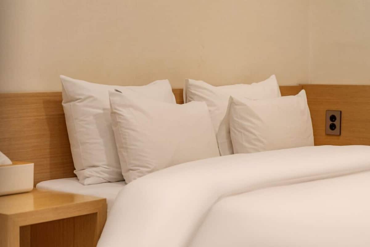 Un letto ben rifatto con lenzuola e cuscini bianchi, una testiera in legno e un comodino con un astuccio di fazzoletti.