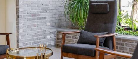 Un área de descanso moderna con pared de ladrillo, una planta grande en maceta y un sillón tapizado en gris oscuro.