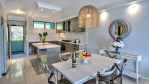 Une cuisine moderne avec des meubles blancs, un coin repas avec une table blanche et des chaises en osier, et un grand miroir au mur.