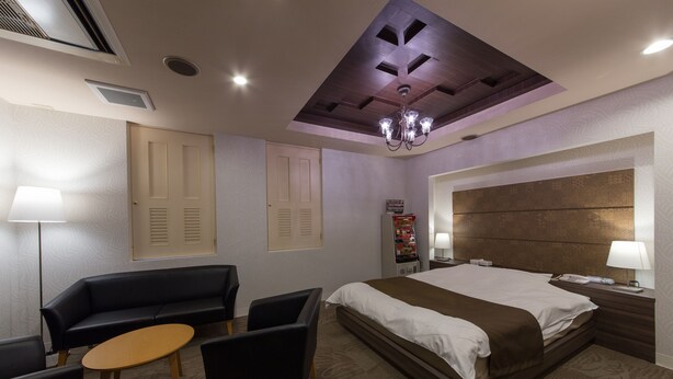 A hotel room with a large bed, a sitting area with a sofa and chairs, a small table, and a nightstand with a lamp. - Hotel Sylph - Adults Only (Wakayama)