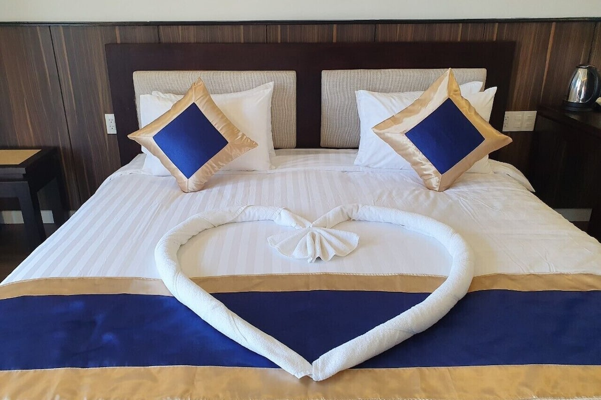 A neatly made bed with a heart-shaped towel arrangement, white bedding, and blue and gold accents.