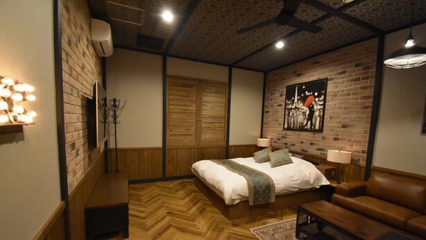 A bedroom with a wooden floor, a bed with a white duvet, a brown leather sofa, a wooden bench, a brick wall, a wooden wardrobe, and a painting on the wall. - Hotel Keypoint - Adults Only (Kai)