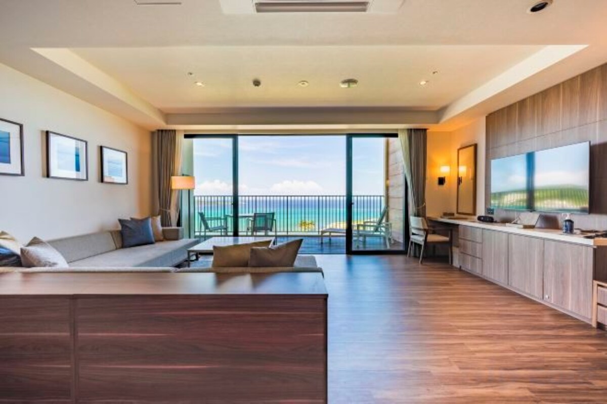 Una habitación de hotel moderna con balcón, sofá, zona de comedor y televisor grande.