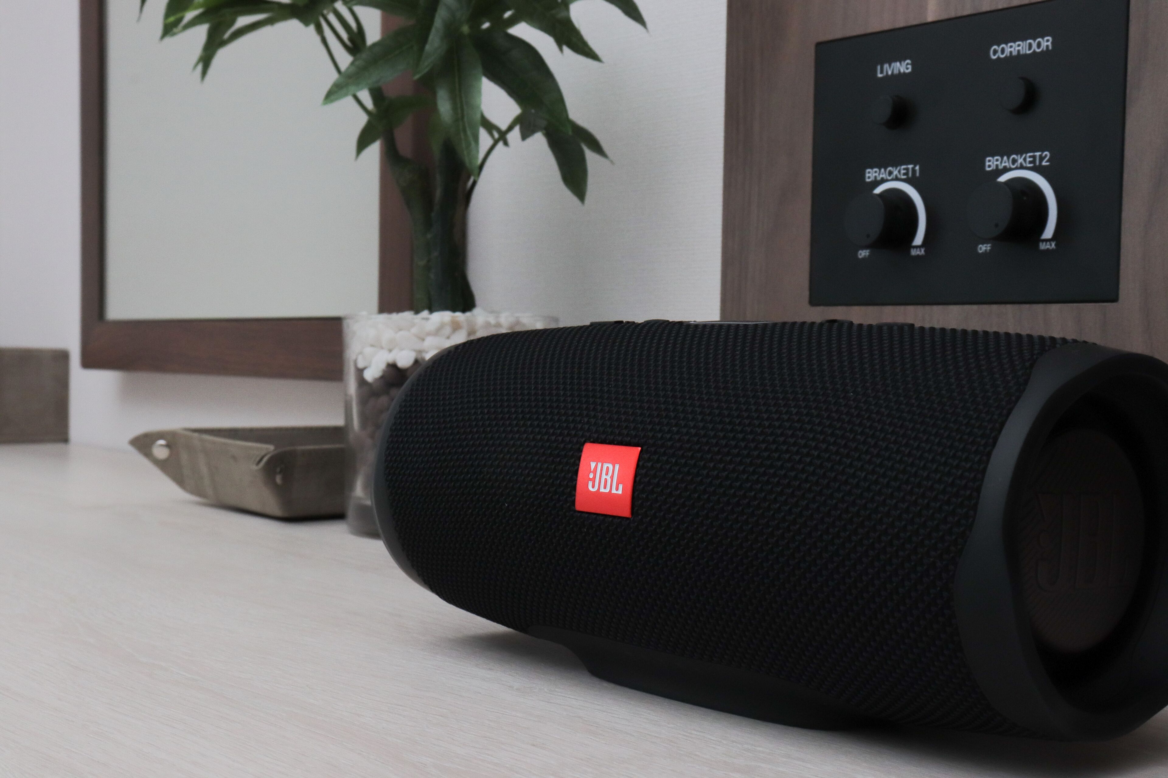 A black JBL speaker on a wooden floor with a plant and a wall-mounted control panel in the background.