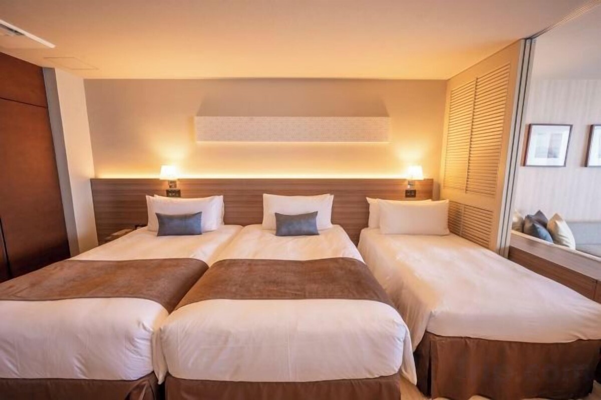 A hotel room with four beds, each with a pillow and a bedside table with a lamp.