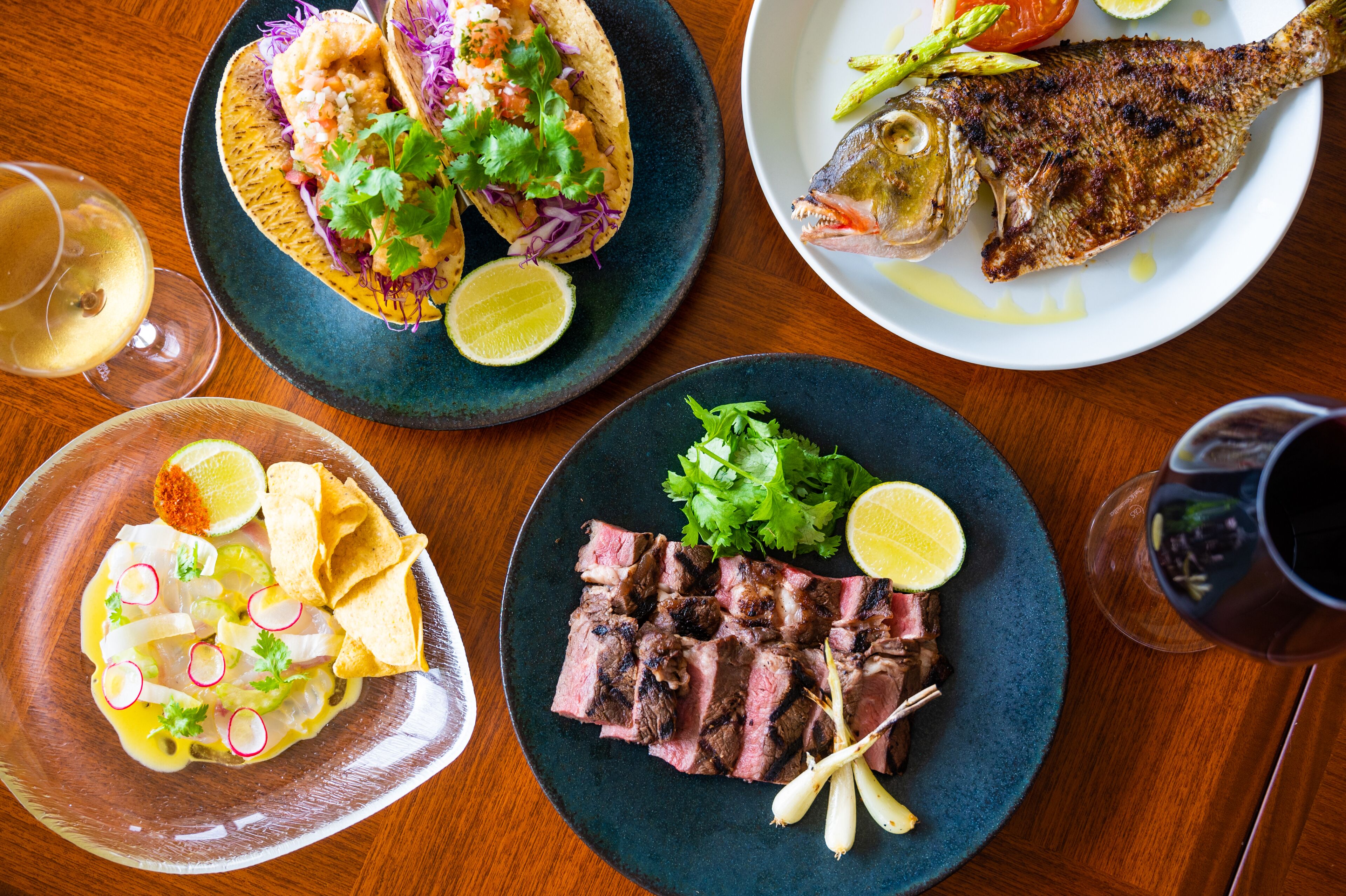 Una mesa con varios platos, incluyendo tacos, pescado a la parrilla, carne y ceviche, acompañados de bebidas.