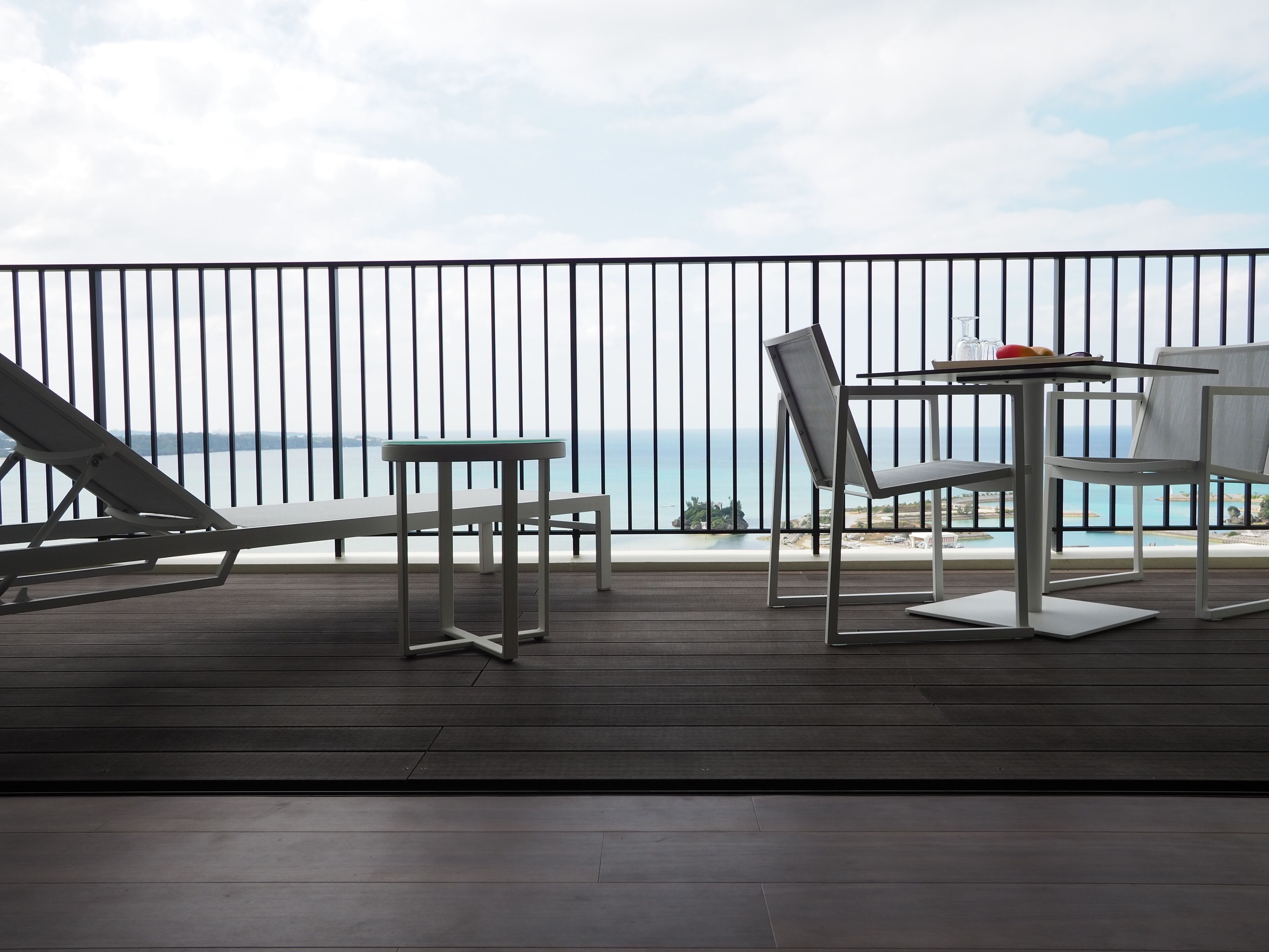 A balcony with a view of the sea, featuring a table, chairs, and a lounge chair.