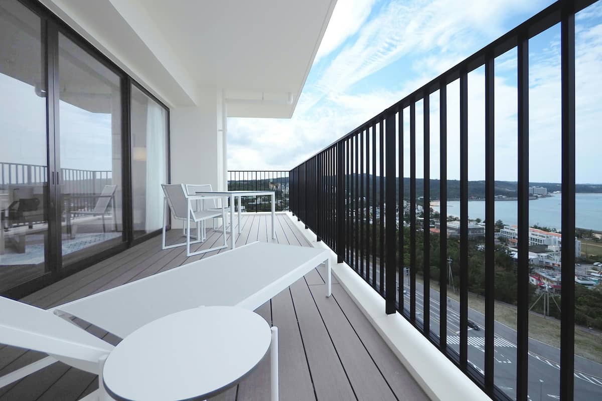 Un balcon avec une table et des chaises blanches, une porte vitrée donnant sur un espace extérieur, et une vue sur une étendue d’eau et des bâtiments.