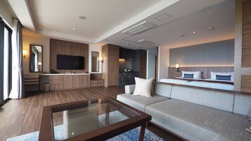 A modern hotel room with a large sofa, a glass coffee table, a flat-screen TV, a kitchen area, and a large window with curtains.