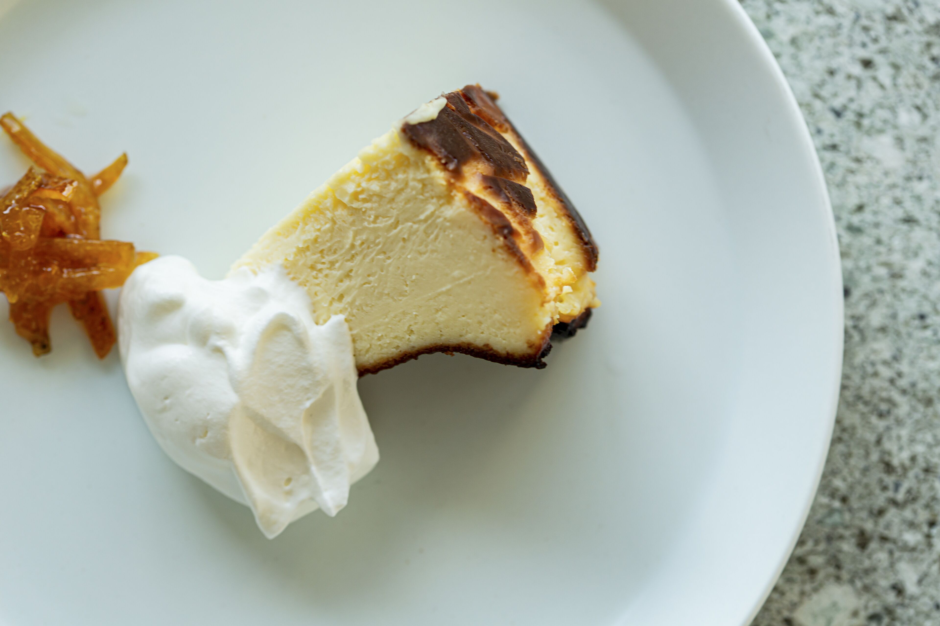 Una porción de pastel de queso con crema batida y fruta caramelizada sobre un plato blanco.