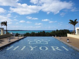 Una piscina infinita con la leyenda 'HIYORI' en el borde, con vista a una playa con palmeras y cielo despejado.