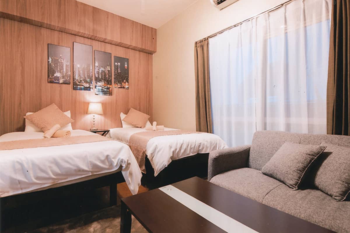 Een hotelkamer met twee bedden, een bank, een houten muur met ingelijste foto’s en een groot raam met gordijnen.