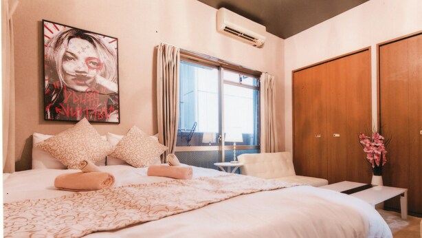 Un dormitorio con una cama grande, una mesita de noche, una silla, una ventana con cortinas, un aire acondicionado instalado en la pared y un cartel en la pared.