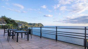 Terraza exterior con mesas y sillas, barandilla y vistas al mar y un paisaje montañoso.