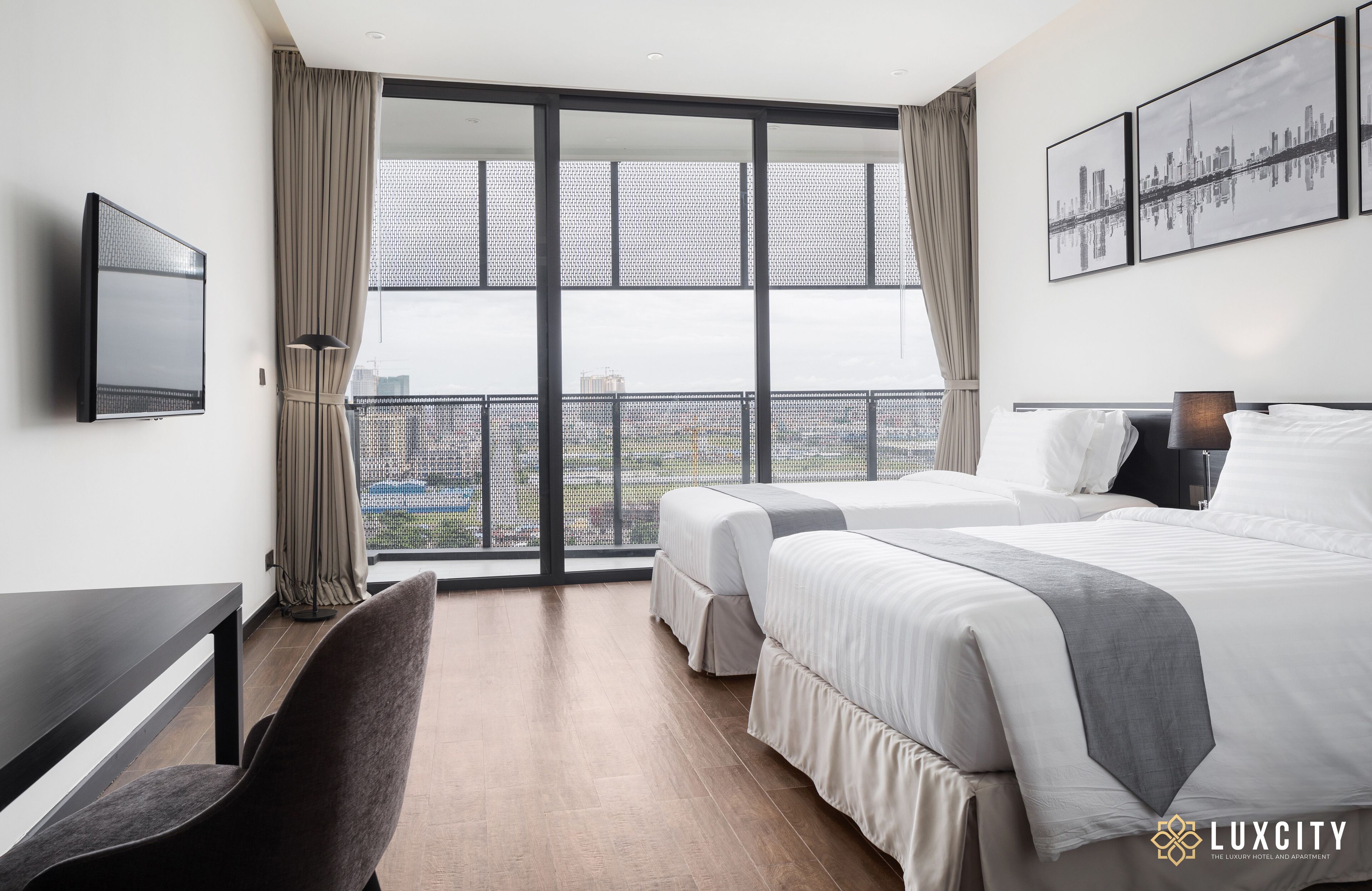 A hotel room with two beds, a desk, a chair, a TV, and a balcony with a city view.