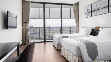 A hotel room with two beds, a desk, a chair, a TV, and a balcony with a city view.