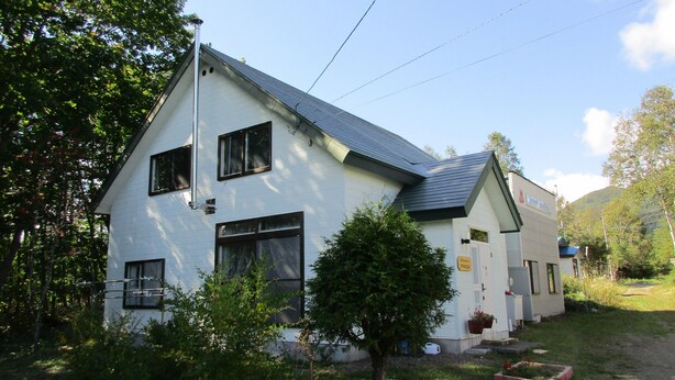 A two-story house with a sloped roof, a small front porch, and a garden area with a tree and flower beds. - Fresh-Air-Inn (Teshikaga)
