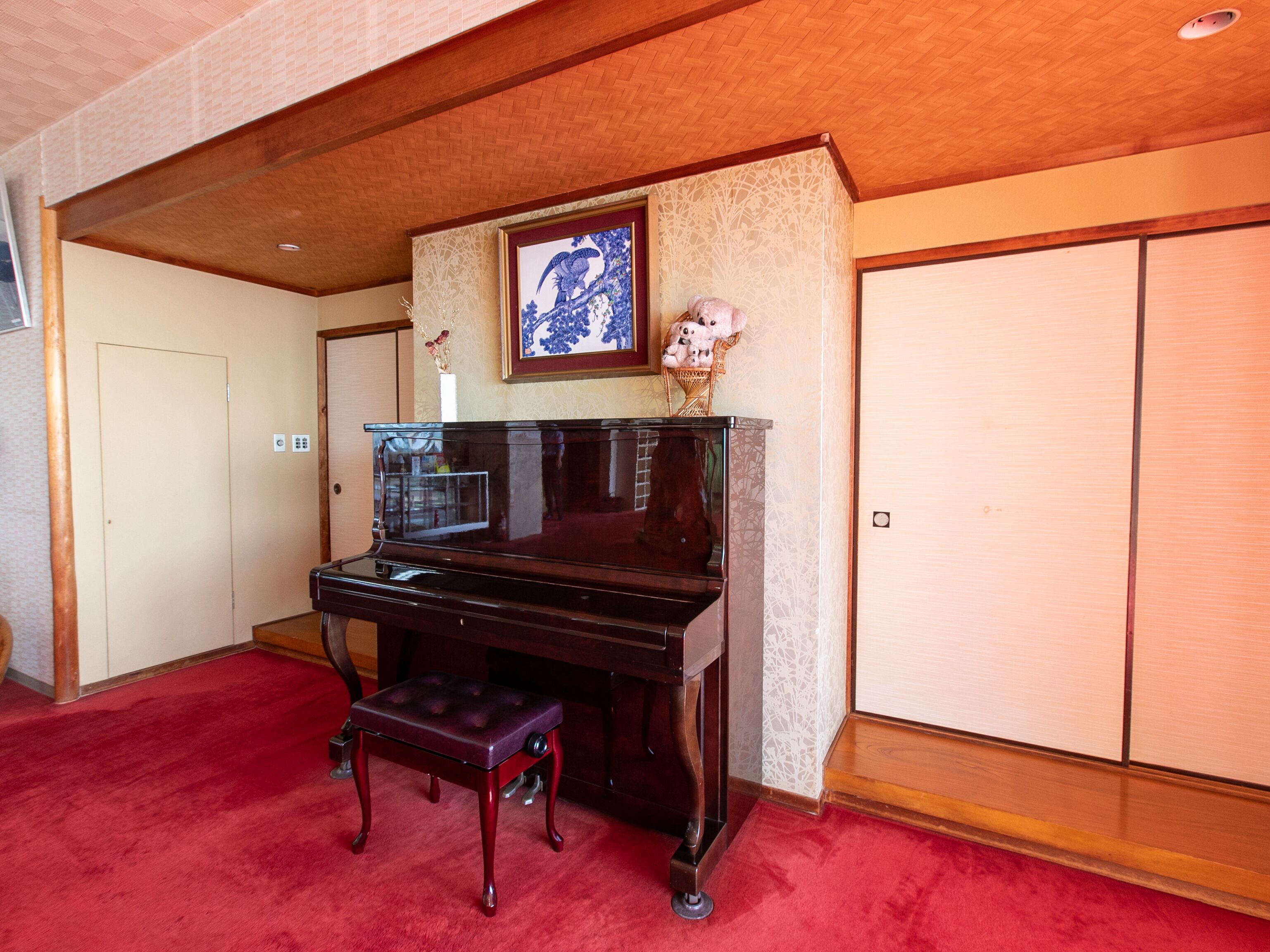 A grand piano in a room with a red carpet, a wooden bench, a framed artwork above the piano, and a door in the background.
