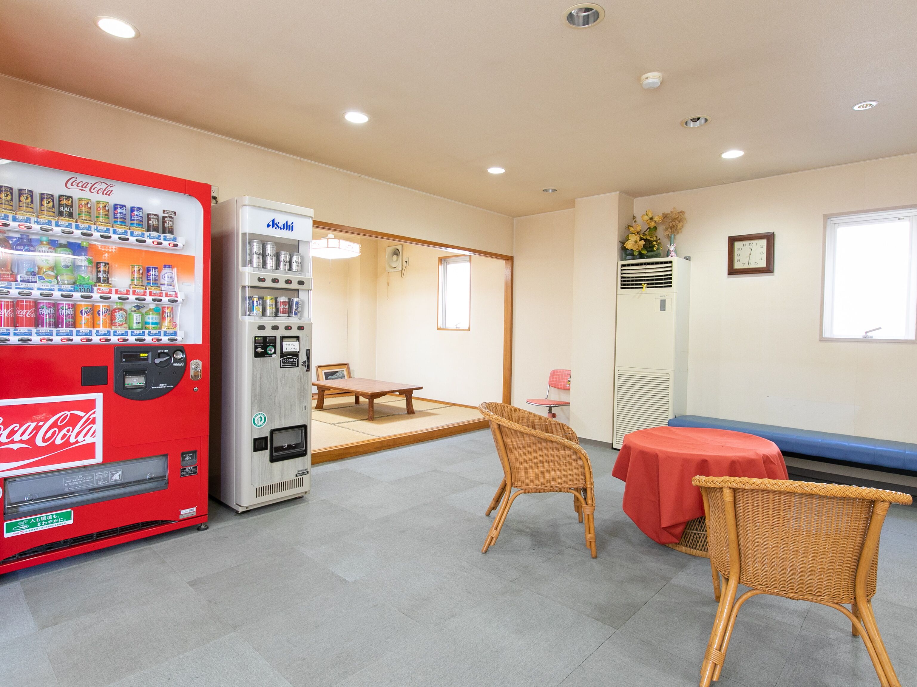 Una habitación con una máquina expendedora roja de Coca-Cola, un refrigerador blanco Asahi, una mesita baja, dos sillas de mimbre y un mantel rojo.