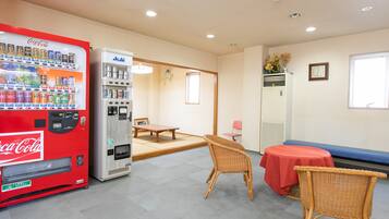 Una habitación con una máquina expendedora roja de Coca-Cola, un refrigerador blanco Asahi, una mesita baja, dos sillas de mimbre y un mantel rojo.