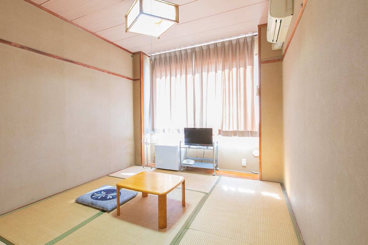 Une pièce de style japonais traditionnel, avec un sol recouvert de tatamis, une petite table en bois, un futon et un téléviseur en arrière-plan.