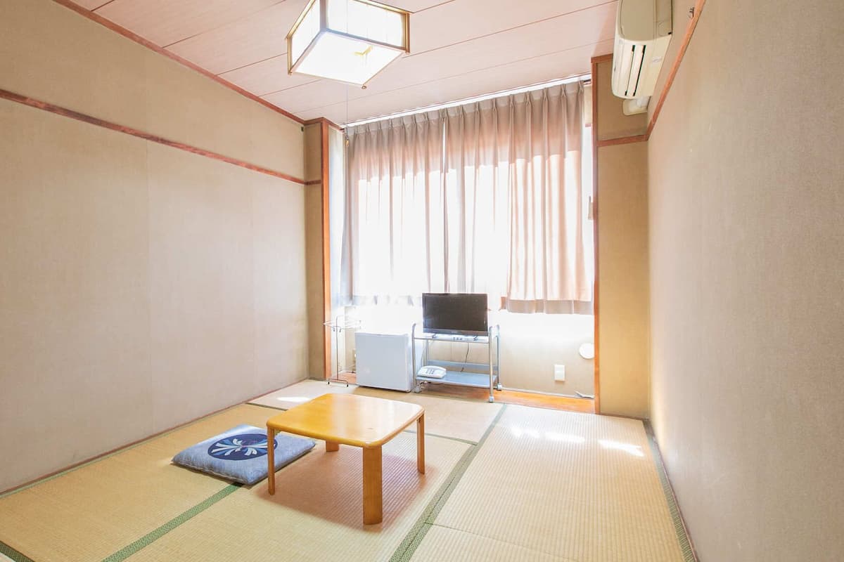 A traditional Japanese room with tatami flooring, a low wooden table, a futon, and a television in the background.