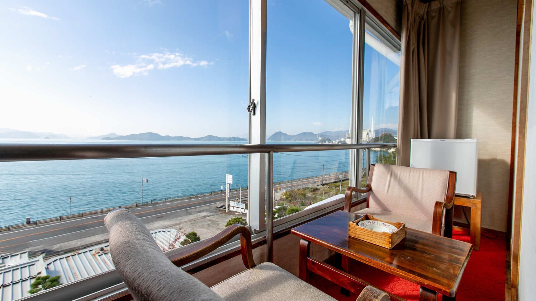 A room with a large window overlooking a body of water and a road, a wooden table with a box, a chair, and a lamp.