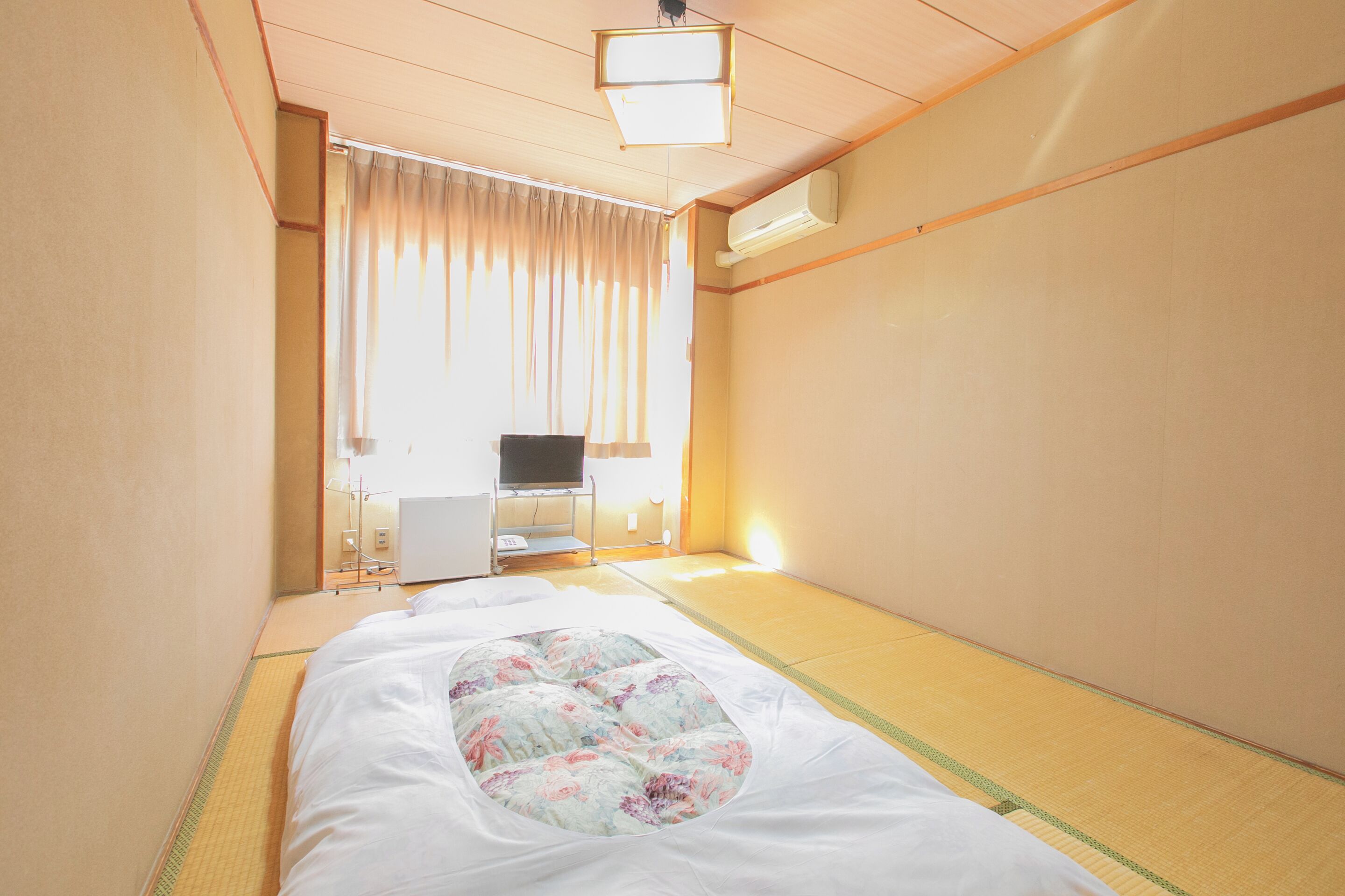 Una tradizionale stanza giapponese con pavimento in tatami, un letto con copripiumino floreale, una televisione e una scrivania con sedia.