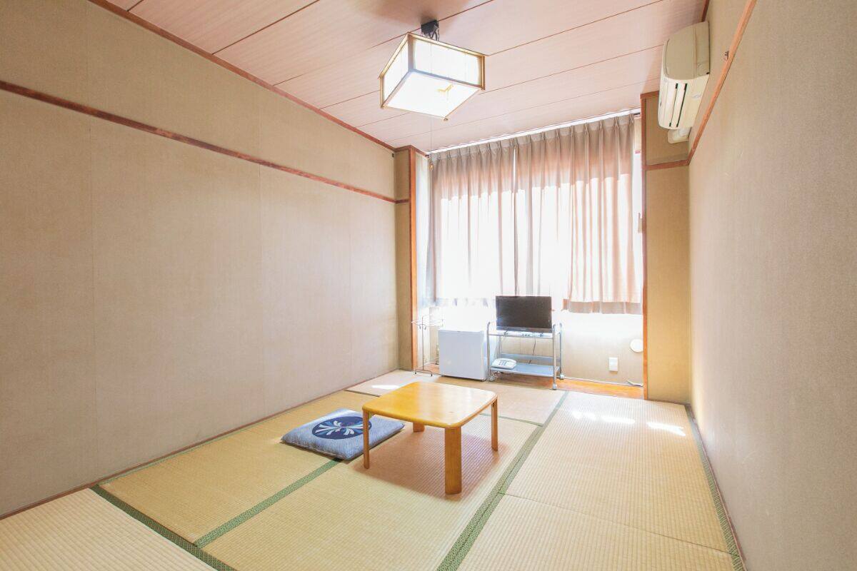 Une pièce de style japonais traditionnel, avec un sol recouvert de tatamis, une petite table en bois, un coussin et un téléviseur en arrière-plan.