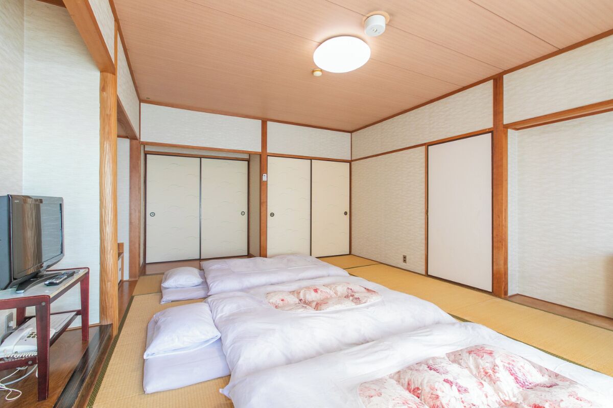 A traditional Japanese-style room with tatami flooring, a bed with white bedding, a television on a wooden stand, and sliding doors.