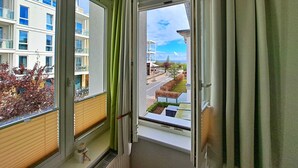 Vista da una finestra con persiane, una tenda e una porta che dà su uno spazio esterno con edifici e cielo sereno.