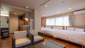 A hotel room with two beds, a kitchenette, a seating area with wicker chairs, and a large window with curtains.