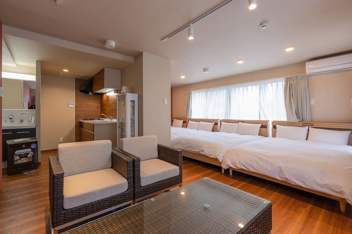 Une chambre d’hôtel avec deux lits, une kitchenette, un coin salon avec des fauteuils en osier et une grande fenêtre avec des rideaux.