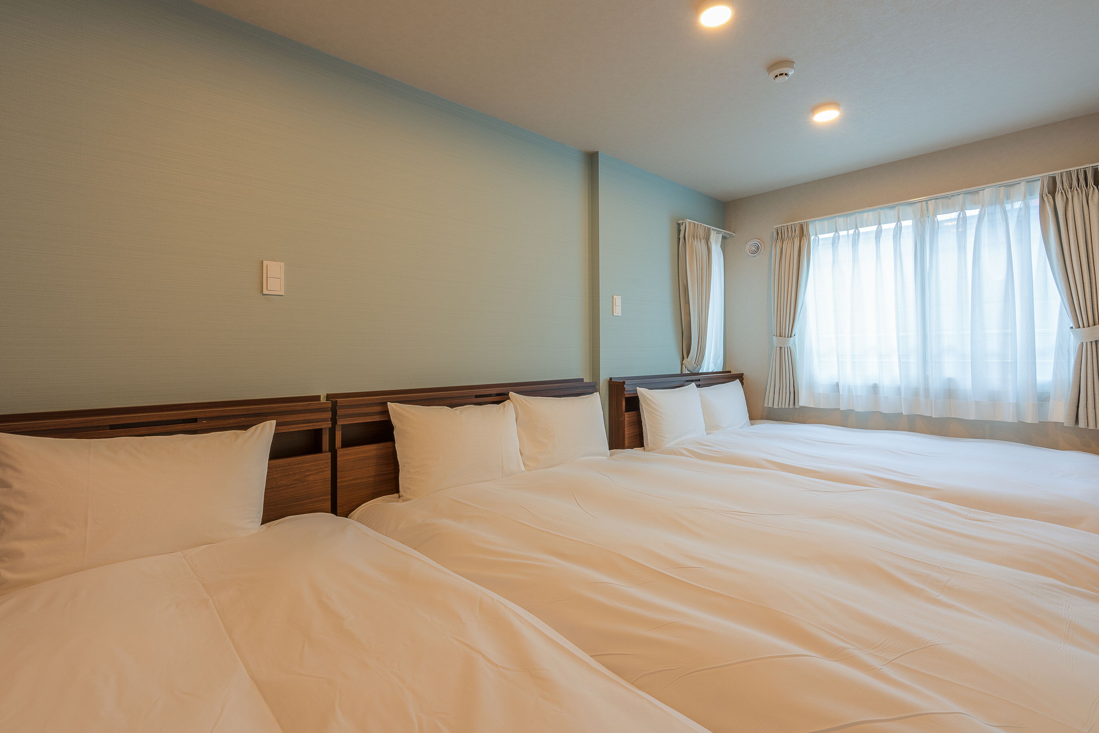 Quarto de hotel com duas camas, cabeceira de madeira, uma janela grande com cortinas e uma luminária no teto.
