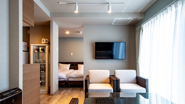 A modern hotel room with a glass dining table, a sofa, a TV mounted on the wall, and a separate bedroom area with a bed and a desk.