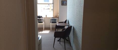 A narrow hallway with a window, a small table with chairs, and a trash bin.