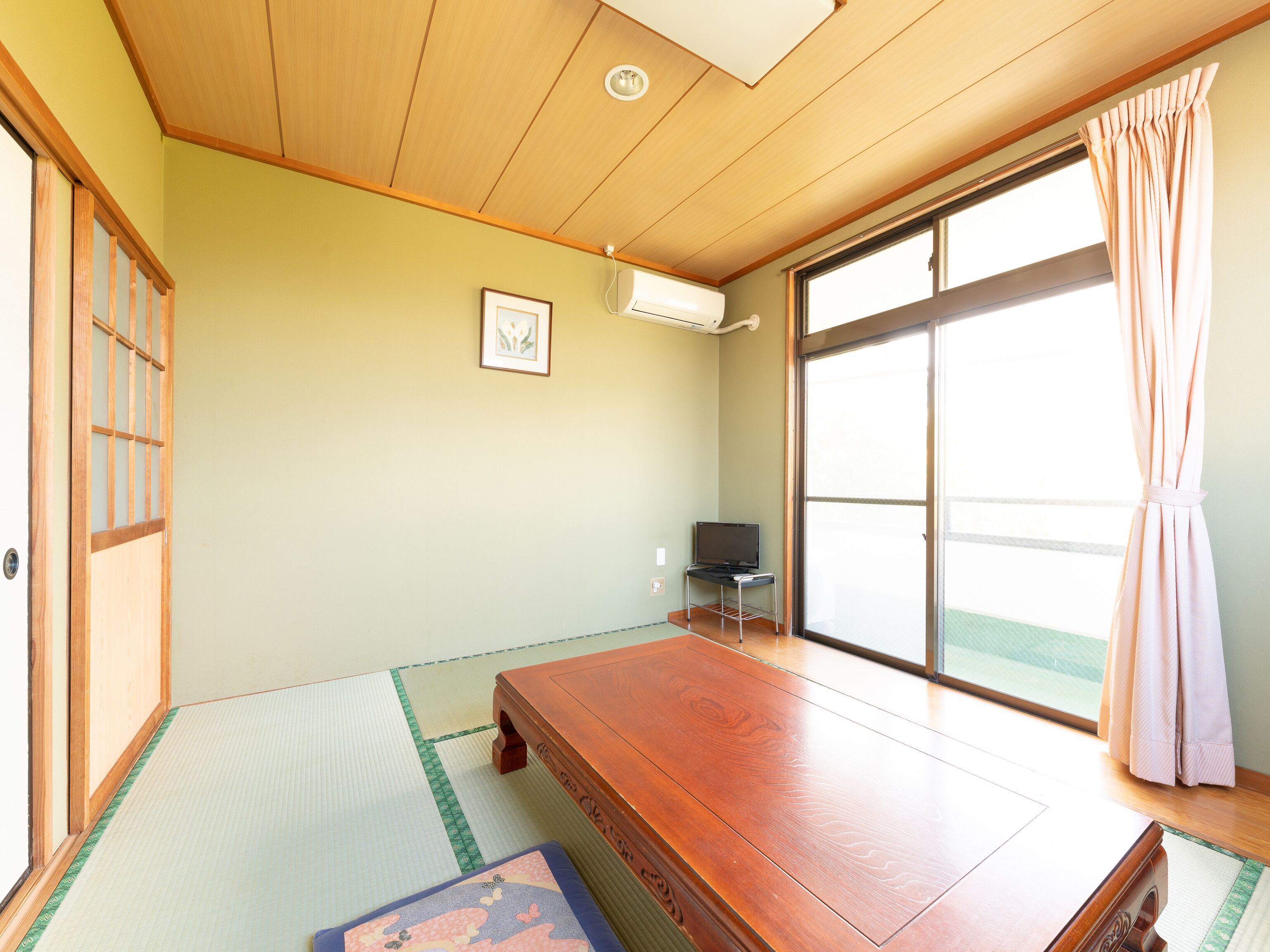 Una habitación japonesa tradicional con suelo de tatami, una mesa de madera, un televisor y una puerta corredera de vidrio con cortinas.