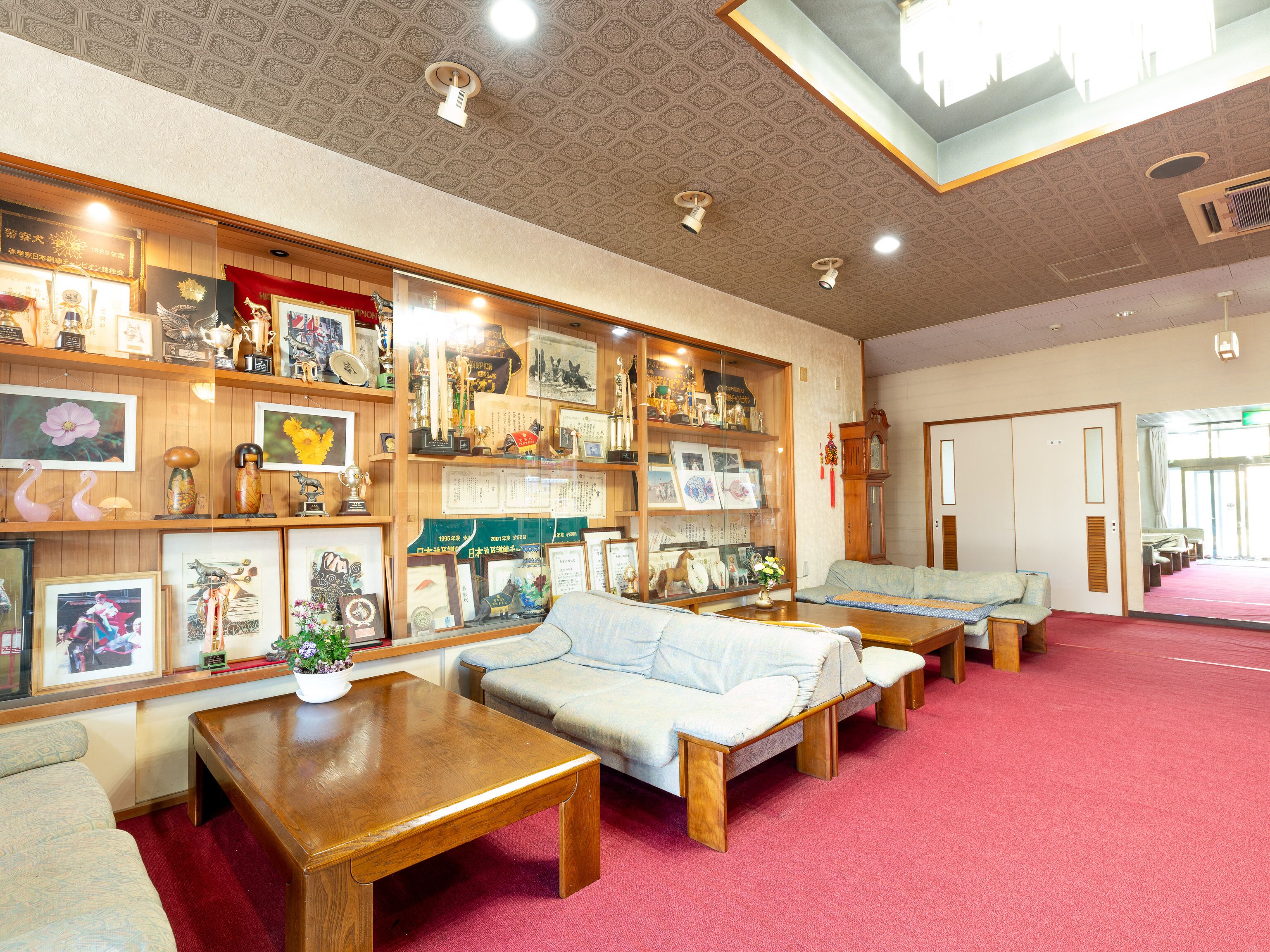 Een kamer met een houten bankje, een houten tafel en een bank. Er staan planken met diverse spullen en een klok aan de muur.