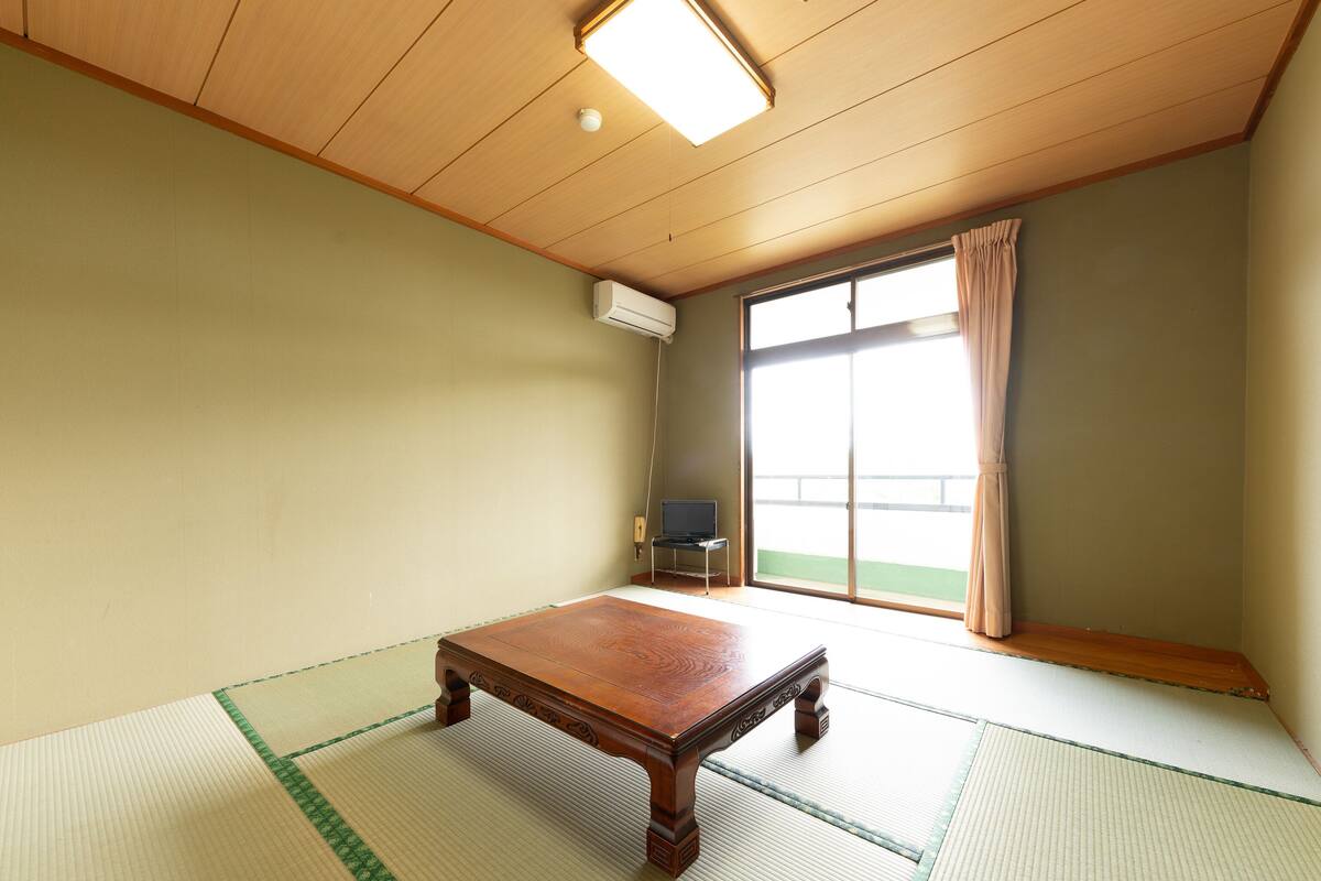 Une pièce avec un sol recouvert de tatamis, une table en bois, un téléviseur et une porte-fenêtre coulissante donnant sur un balcon.