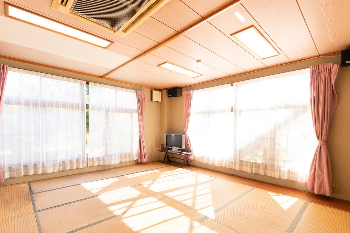 Une pièce avec un sol recouvert de tatamis, de grandes fenêtres avec des rideaux légers, un téléviseur posé sur une petite table et un ventilateur de plafond.