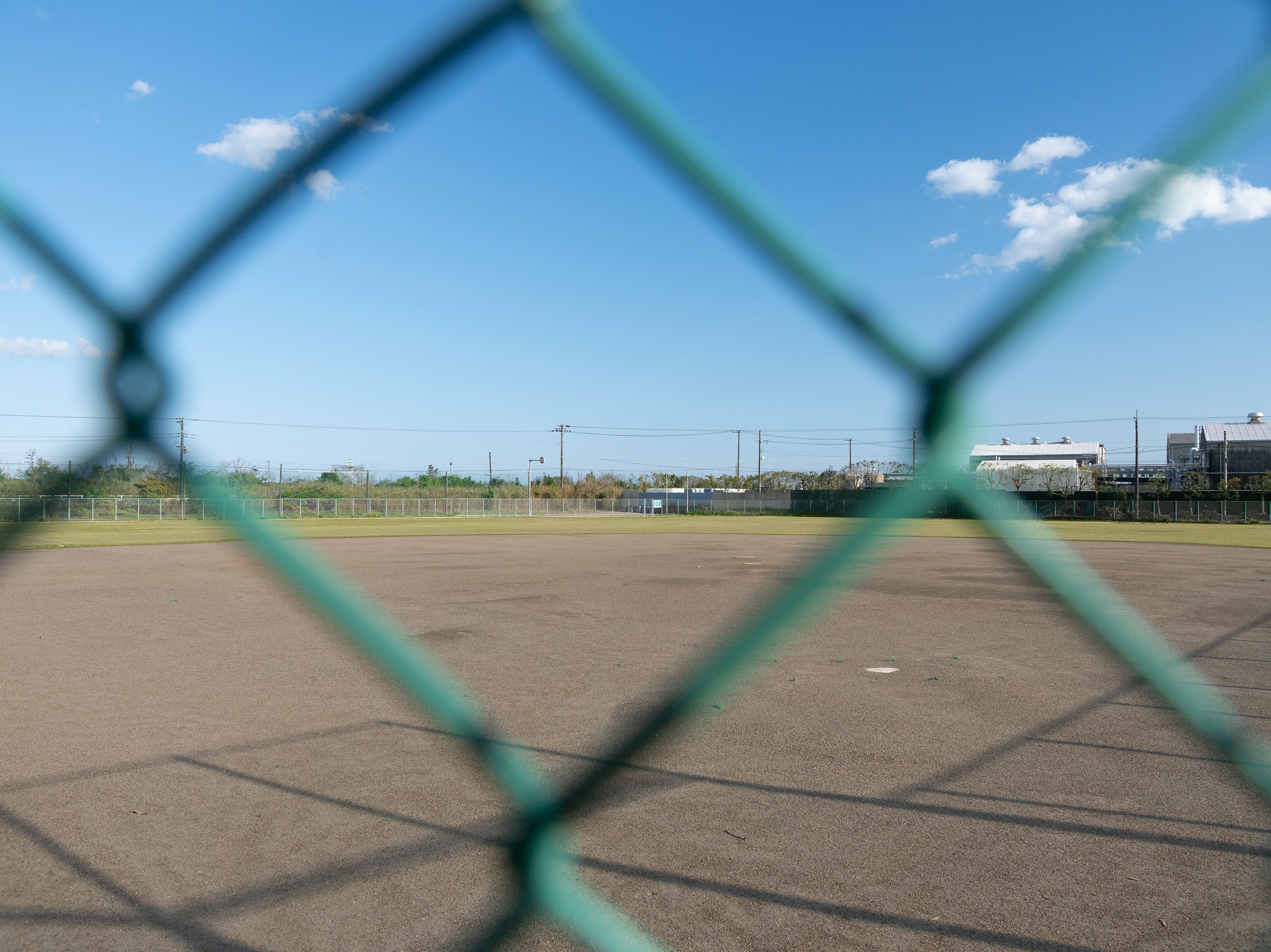 Un campo deportivo con una cerca de malla metálica, cielo despejado y edificios industriales al fondo.