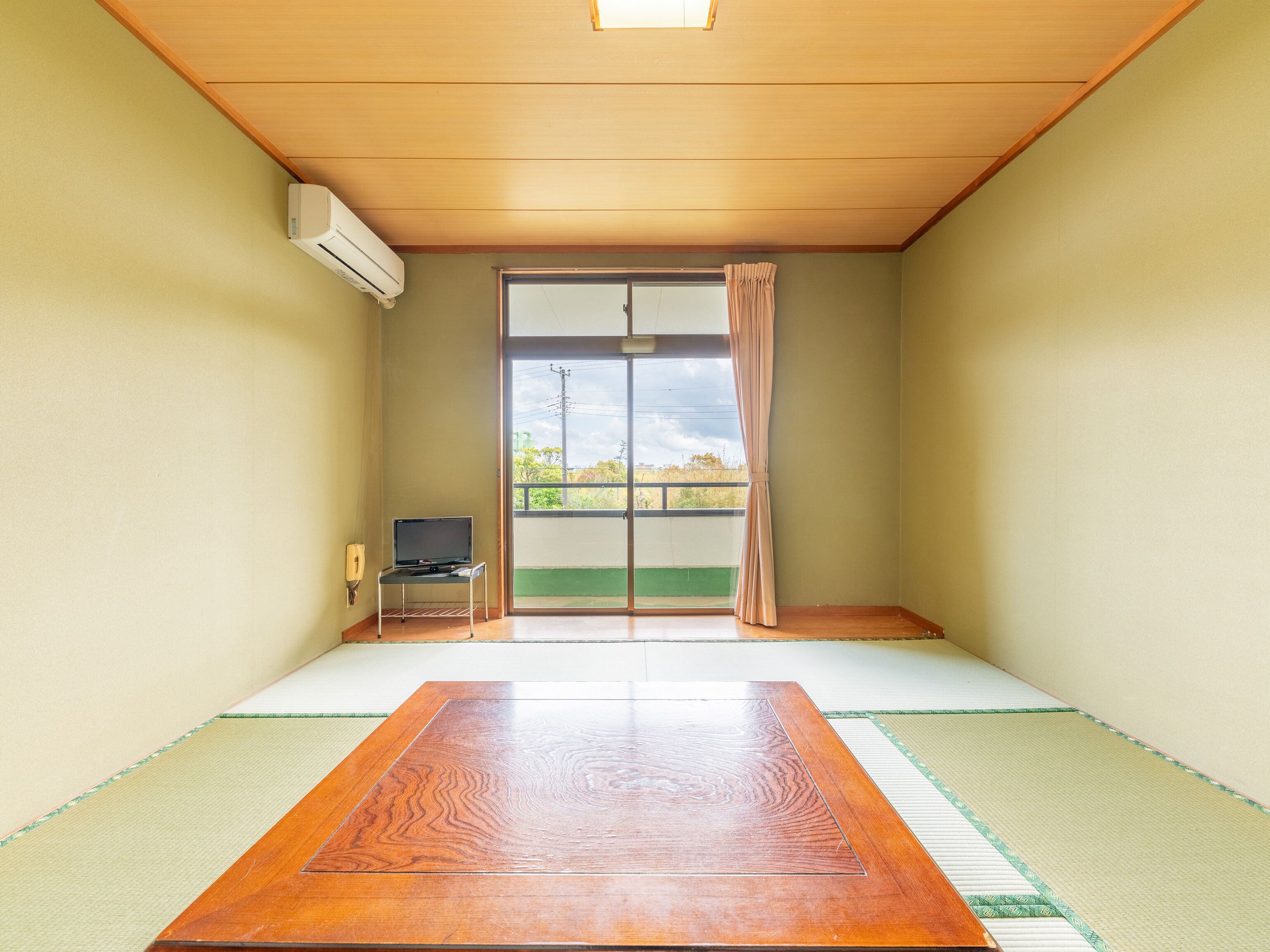 Une pièce avec un sol recouvert de tatamis, une table en bois, un téléviseur posé sur un meuble et une porte coulissante donnant sur un balcon.